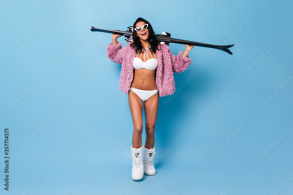 Full length view of happy girl in bikini holding skis. Studio shot of charming slim woman in swimsuit and jacket.