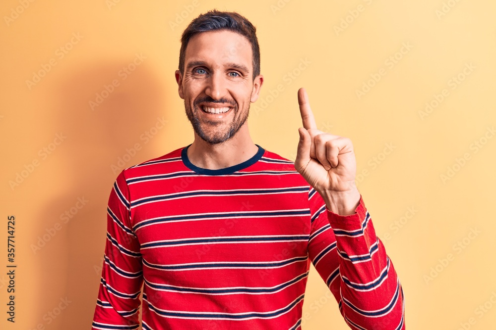 Young handsome man wearing casual striped t-shirt standing over isolated yellow background smiling with an idea or question pointing finger up with happy face, number one Young handsome man wearing casual striped t-shirt standing over isolated yellow background smiling with an idea or question pointing finger up with happy face, number one