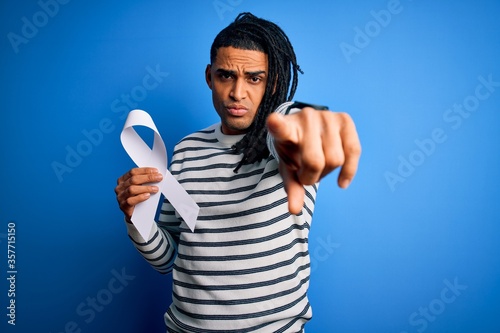 Young african american man with dreadlocks holding white ribbon over blue background pointing with finger to the camera and to you, hand sign, positive and confident gesture from the front