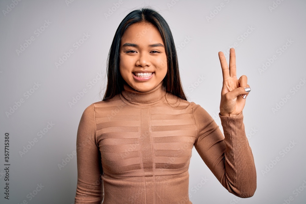 Young beautiful asian girl wearing casual turtleneck sweater standing over white background smiling with happy face winking at the camera doing victory sign. Number two. Young beautiful asian girl wearing casual turtleneck sweater standing over white background smiling with happy face winking at the camera doing victory sign. Number two.