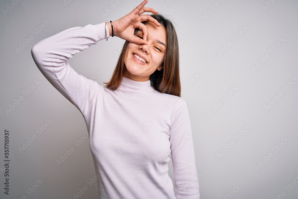 Young beautiful brunette girl wearing casual sweater standing over isolated white background doing ok gesture with hand smiling, eye looking through fingers with happy face. Young beautiful brunette girl wearing casual sweater standing over isolated white background doing ok gesture with hand smiling, eye looking through fingers with happy face.