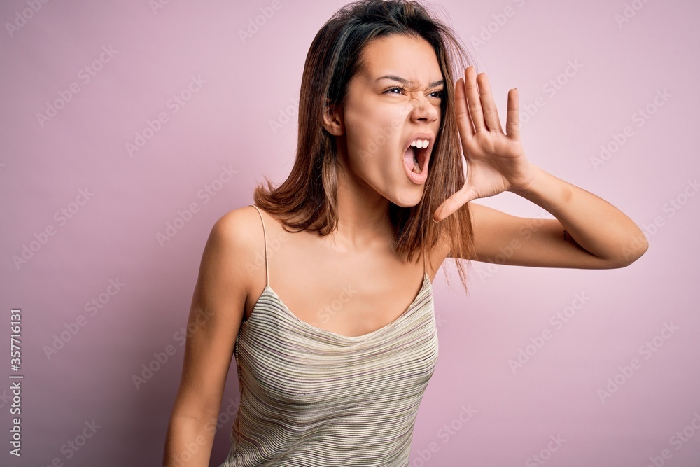 Young beautiful brunette girl wearing casual striped t-shirt over isolated pink background shouting and screaming loud to side with hand on mouth. Communication concept. Young beautiful brunette girl wearing casual striped t-shirt over isolated pink background shouting and screaming loud to side with hand on mouth. Communication concept.