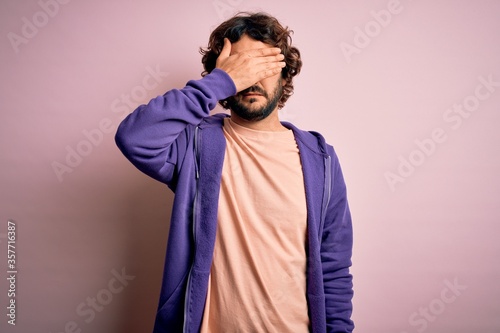 Young handsome sporty man with beard wearing casual sweatshirt over pink background covering eyes with hand, looking serious and sad. Sightless, hiding and rejection concept