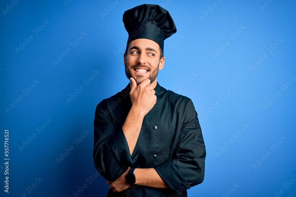 Young handsome chef man with beard wearing cooker uniform and hat over blue background with hand on chin thinking about question, pensive expression. Smiling with thoughtful face. Doubt concept. Young handsome chef man with beard wearing cooker uniform and hat over blue background with hand on chin thinking about question, pensive expression. Smiling with thoughtful face. Doubt concept.