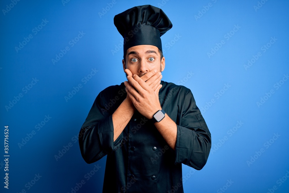 Young handsome chef man with beard wearing cooker uniform and hat over blue background shocked covering mouth with hands for mistake. Secret concept. Young handsome chef man with beard wearing cooker uniform and hat over blue background shocked covering mouth with hands for mistake. Secret concept.