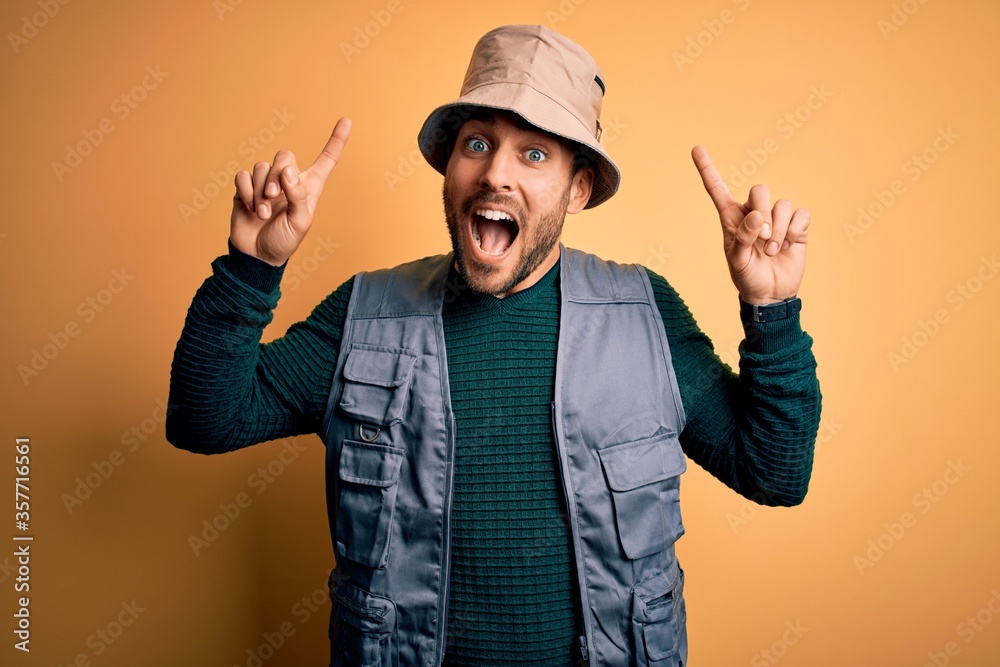Handsome tourist man with beard on vacation wearing explorer hat over yellow background smiling amazed and surprised and pointing up with fingers and raised arms.
