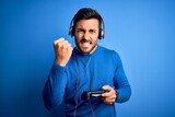 Young handsome gamer man with beard playing video game using joystick and headphones annoyed and frustrated shouting with anger, crazy and yelling with raised hand, anger concept