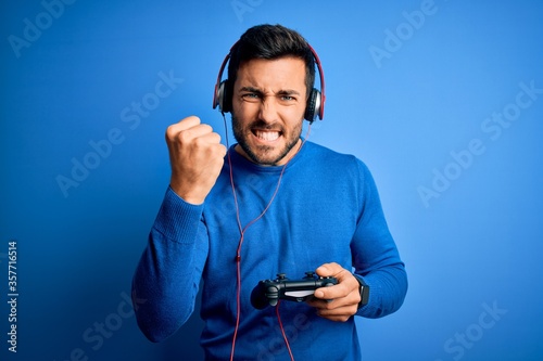 Young handsome gamer man with beard playing video game using joystick and headphones annoyed and frustrated shouting with anger, crazy and yelling with raised hand, anger concept photo