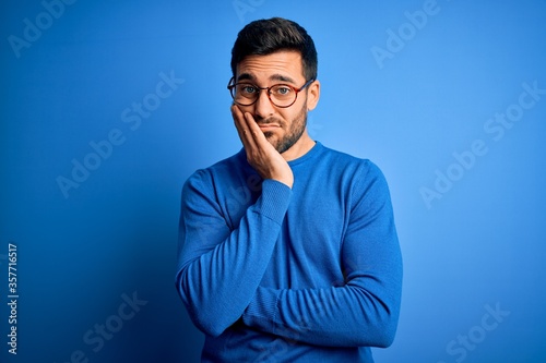 Young handsome man with beard wearing casual sweater and glasses over blue background thinking looking tired and bored with depression problems with crossed arms.