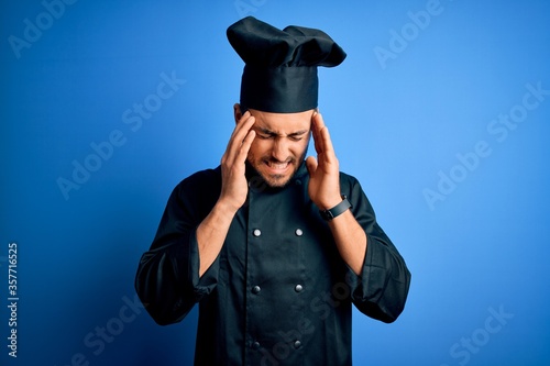 Young handsome chef man with beard wearing cooker uniform and hat over blue background with hand on head for pain in head because stress. Suffering migraine.