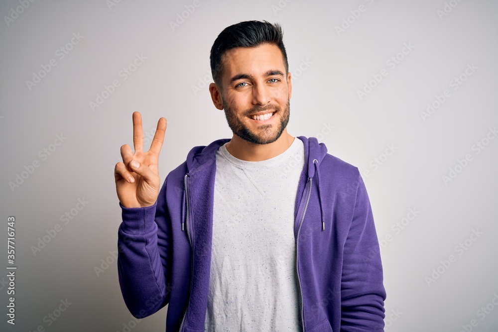 Young handsome man wearing purple sweatshirt standing over isolated white background smiling with happy face winking at the camera doing victory sign. Number two.