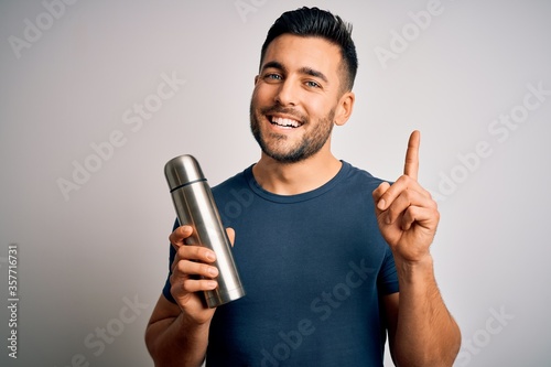 Young handsome tourist man drinking thermo with water over isolated white background surprised with an idea or question pointing finger with happy face, number one