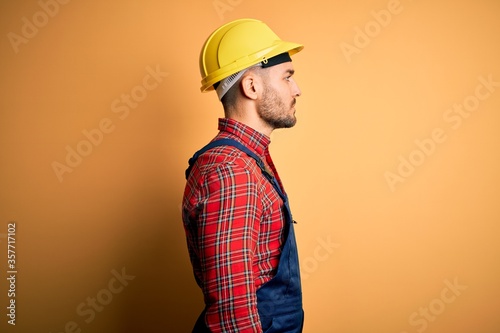 Young builder man wearing construction uniform and safety helmet over yellow isolated background looking to side  relax profile pose with natural face with confident smile.
