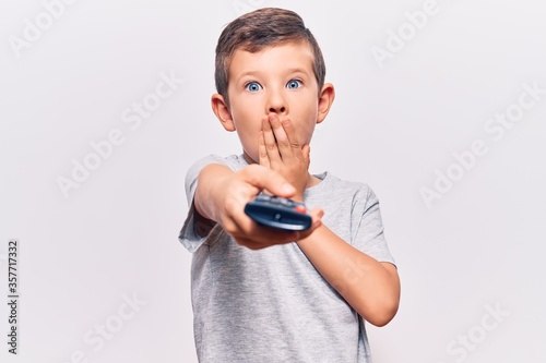 Cute blond kid holding television remote control covering mouth with hand, shocked and afraid for mistake. surprised expression