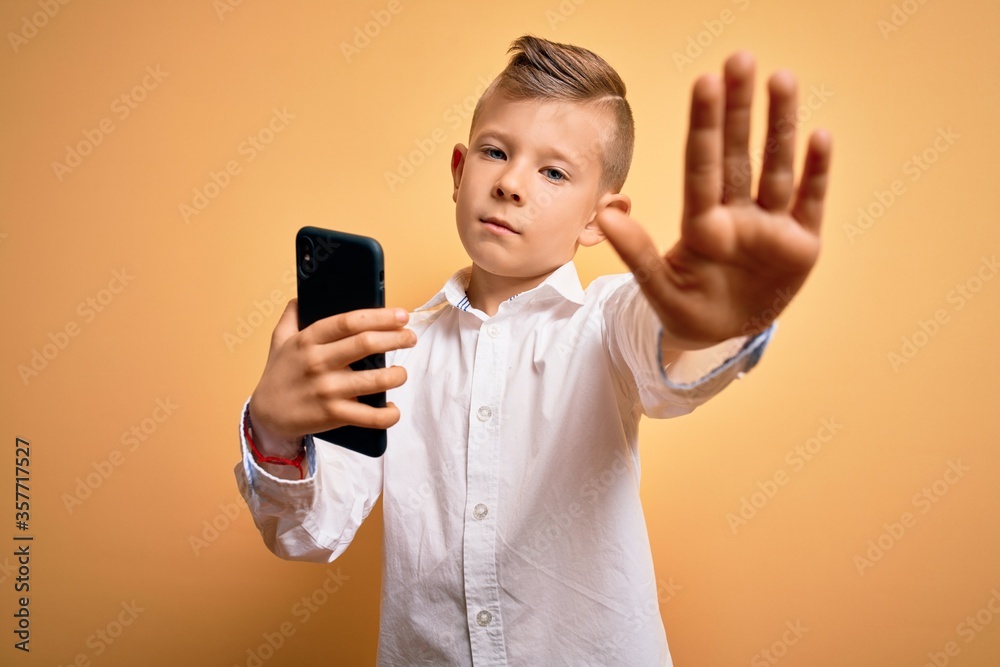 Young little caucasian kid using smartphone and looking at the screen of the phone with open hand doing stop sign with serious and confident expression, defense gesture Young little caucasian kid using smartphone and looking at the screen of the phone with open hand doing stop sign with serious and confident expression, defense gesture