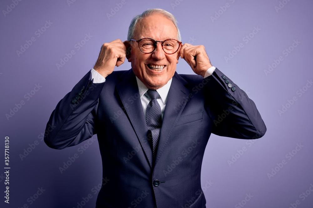 Grey haired senior business man wearing glasses and elegant suit and tie over purple background Smiling pulling ears with fingers, funny gesture. Audition problem