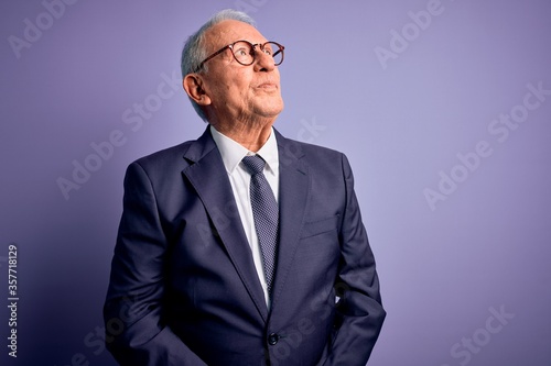 Grey haired senior business man wearing glasses and elegant suit and tie over purple background smiling looking to the side and staring away thinking.
