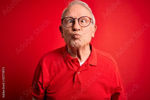 Grey haired senior man wearing glasses and casual t-shirt over red background making fish face with lips, crazy and comical gesture. Funny expression. © Krakenimages.com