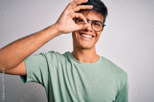 Young handsome man wearing casual t-shirt and glasses over isolated white background doing ok gesture with hand smiling, eye looking through fingers with happy face. © Krakenimages.com
