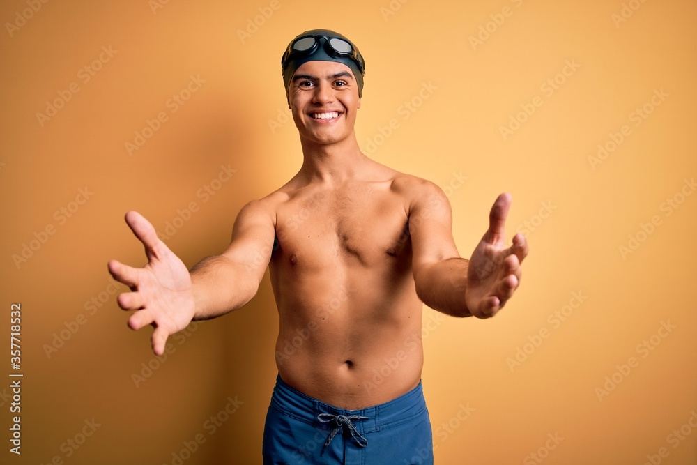 Young handsome man shirtless wearing swimsuit and swim cap over isolated yellow background looking at the camera smiling with open arms for hug. Cheerful expression embracing happiness.