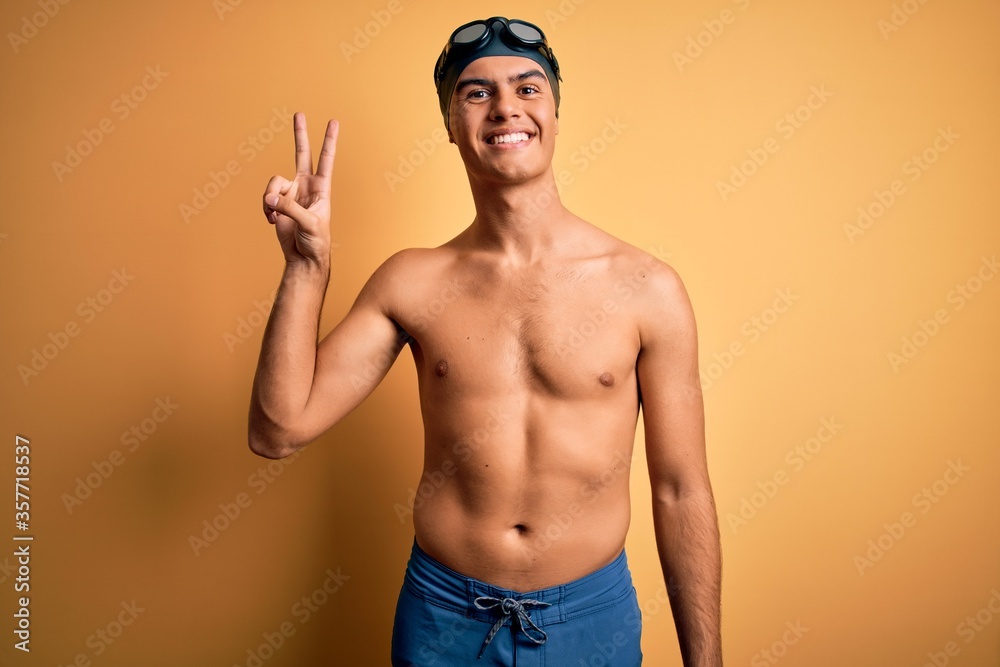 Young handsome man shirtless wearing swimsuit and swim cap over isolated yellow background smiling looking to the camera showing fingers doing victory sign. Number two.