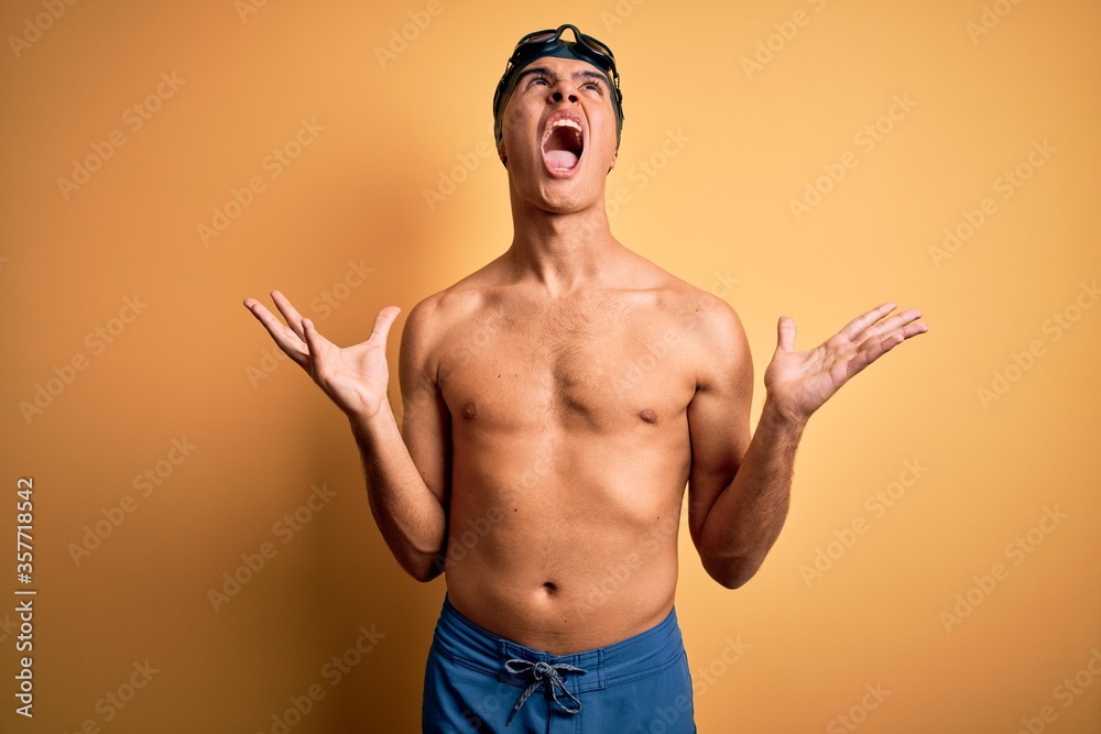 Young handsome man shirtless wearing swimsuit and swim cap over isolated yellow background crazy and mad shouting and yelling with aggressive expression and arms raised. Frustration concept.