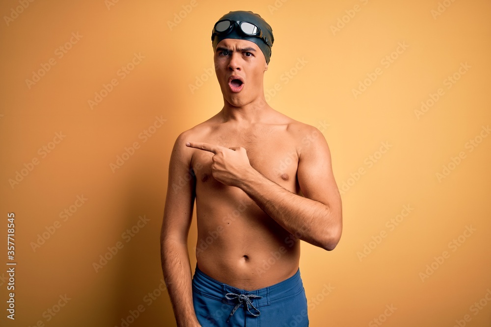 Young handsome man shirtless wearing swimsuit and swim cap over isolated yellow background Surprised pointing with finger to the side, open mouth amazed expression.