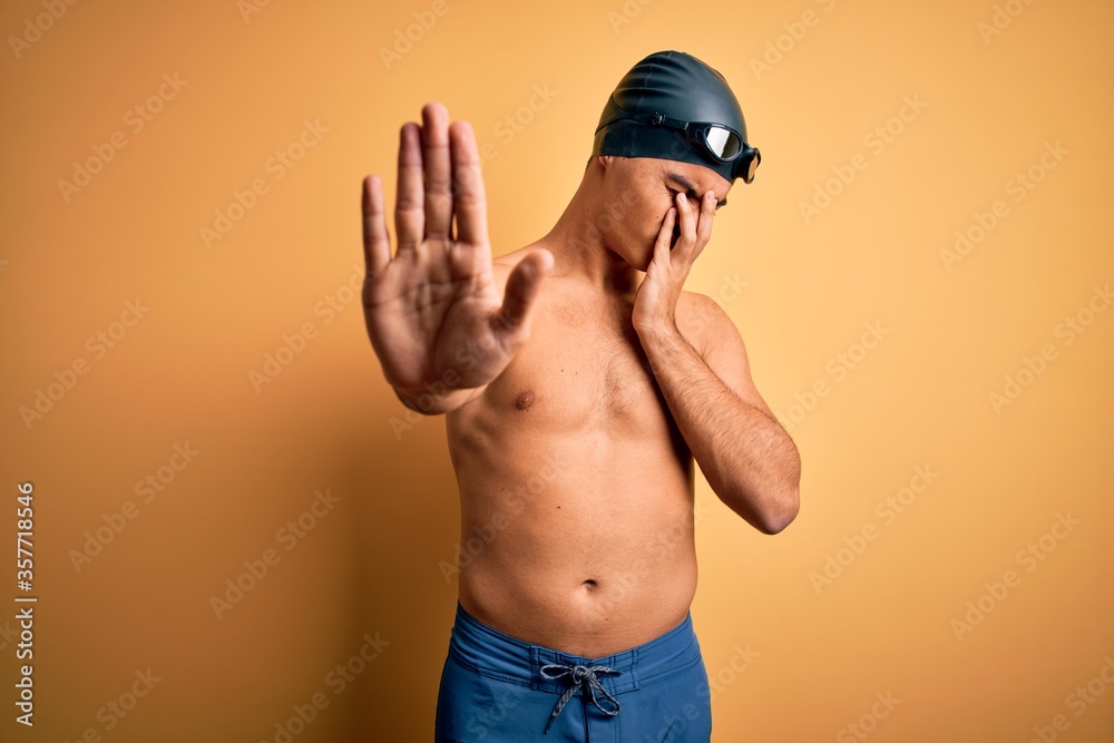 Young handsome man shirtless wearing swimsuit and swim cap over isolated yellow background covering eyes with hands and doing stop gesture with sad and fear expression. Embarrassed and negative