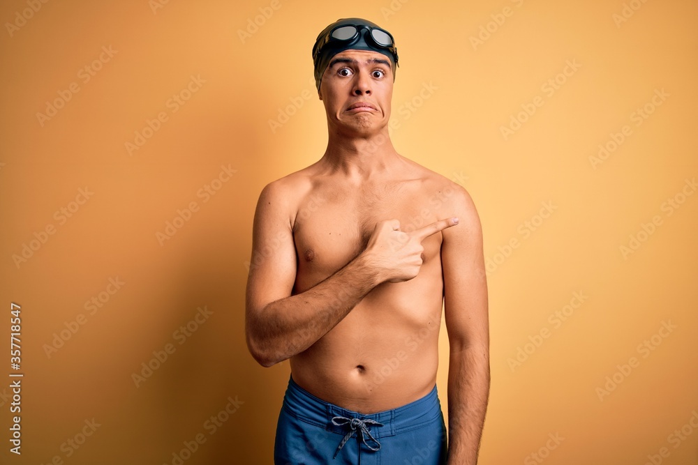 Young handsome man shirtless wearing swimsuit and swim cap over isolated yellow background Pointing aside worried and nervous with forefinger, concerned and surprised expression