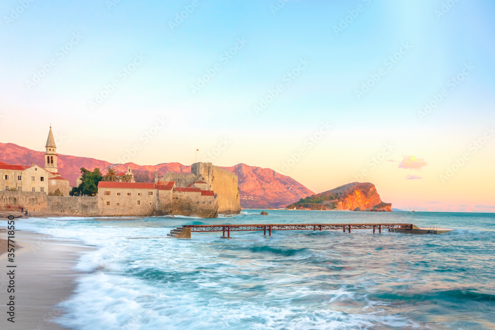 Waves rolling onto the beach and an abandoned pier. Old city in the sunset light. Summer seascape of Montenegro.