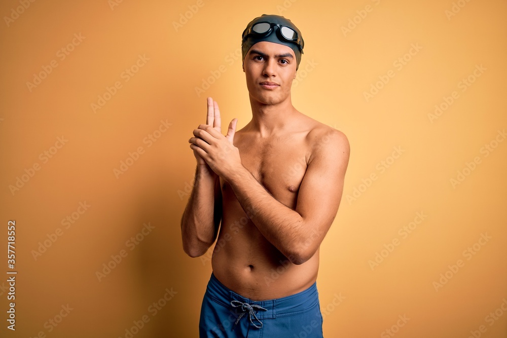 Young handsome man shirtless wearing swimsuit and swim cap over isolated yellow background Holding symbolic gun with hand gesture, playing killing shooting weapons, angry face