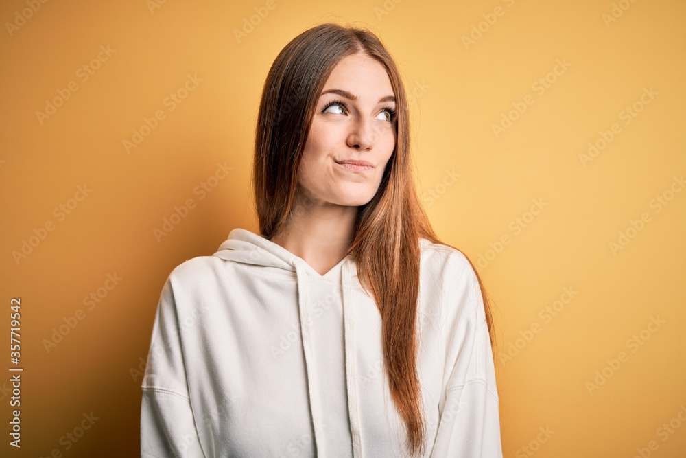 Young beautiful redhead sporty woman wearing sweatshirt over isolated yellow background smiling looking to the side and staring away thinking. Young beautiful redhead sporty woman wearing sweatshirt over isolated yellow background smiling looking to the side and staring away thinking.