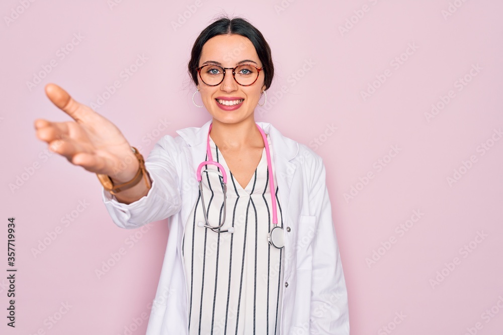 Beautiful doctor woman with blue eyes wearing coat and stethoscope over pink background smiling friendly offering handshake as greeting and welcoming. Successful business. Beautiful doctor woman with blue eyes wearing coat and stethoscope over pink background smiling friendly offering handshake as greeting and welcoming. Successful business.