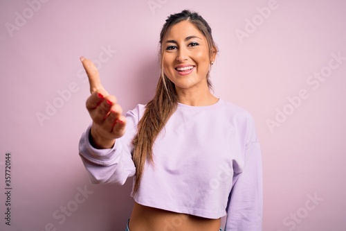 Young beautiful sport woman wearing sweatshirt over pink isolated background smiling friendly offering handshake as greeting and welcoming. Successful business.