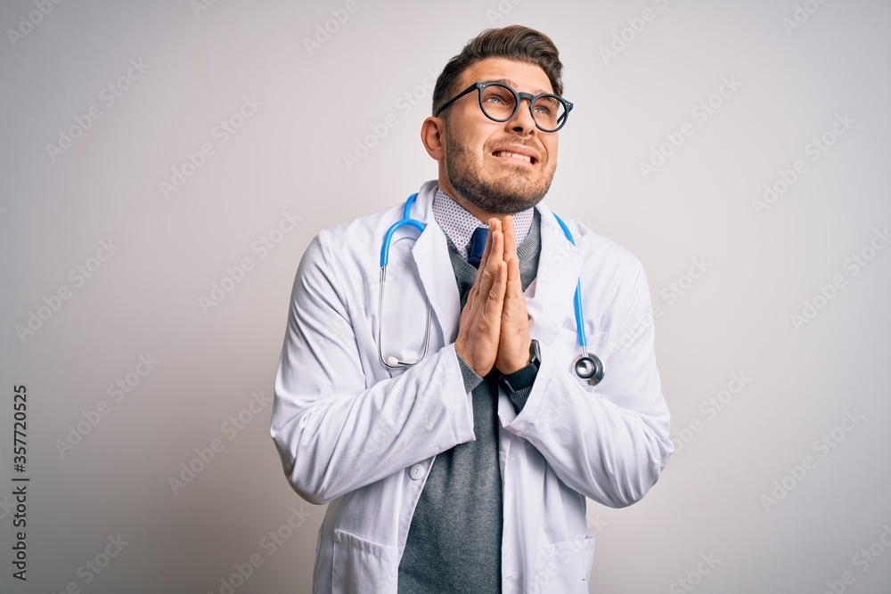 Young doctor man with blue eyes wearing medical coat and stethoscope over isolated background begging and praying with hands together with hope expression on face very emotional and worried. Begging. Young doctor man with blue eyes wearing medical coat and stethoscope over isolated background begging and praying with hands together with hope expression on face very emotional and worried. Begging.