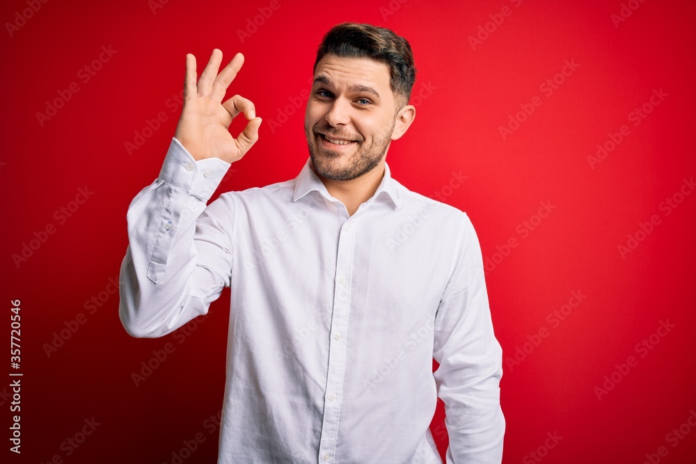 Young business man with blue eyes wearing elegant shirt standing over red isolated background smiling positive doing ok sign with hand and fingers. Successful expression. Young business man with blue eyes wearing elegant shirt standing over red isolated background smiling positive doing ok sign with hand and fingers. Successful expression.