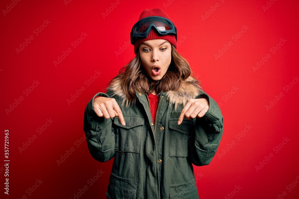 Young blonde girl wearing ski glasses and winter coat for ski weather over red background Pointing down with fingers showing advertisement, surprised face and open mouth Young blonde girl wearing ski glasses and winter coat for ski weather over red background Pointing down with fingers showing advertisement, surprised face and open mouth