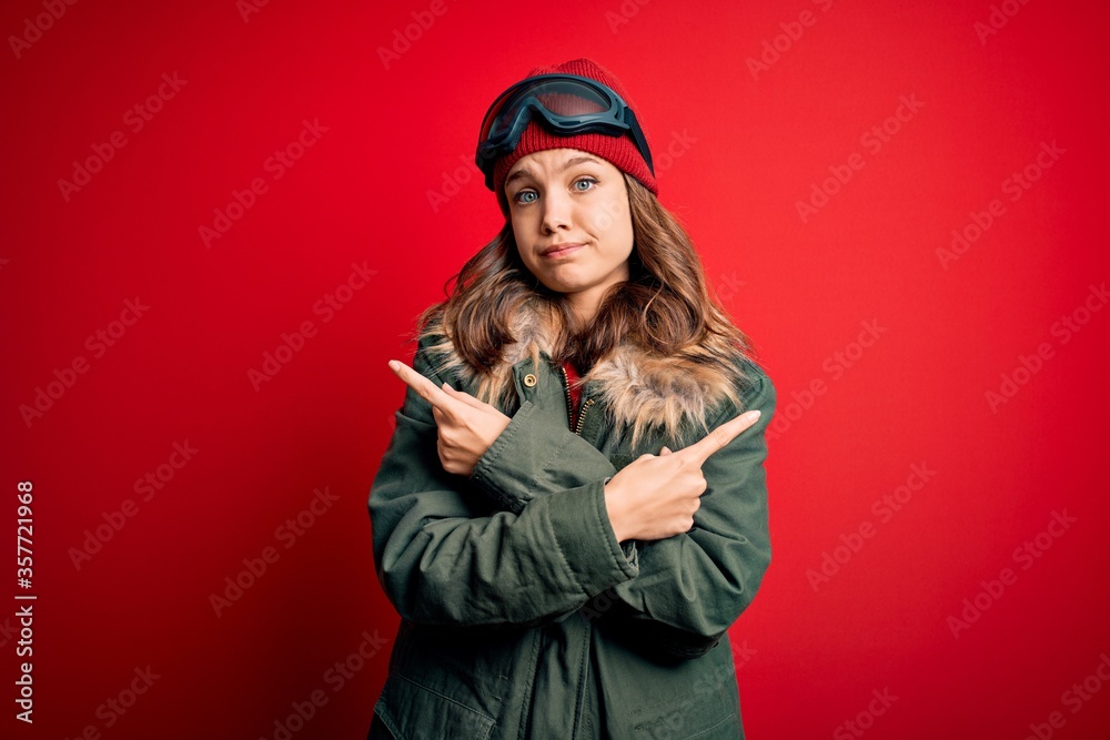 Young blonde girl wearing ski glasses and winter coat for ski weather over red background Pointing to both sides with fingers, different direction disagree Young blonde girl wearing ski glasses and winter coat for ski weather over red background Pointing to both sides with fingers, different direction disagree