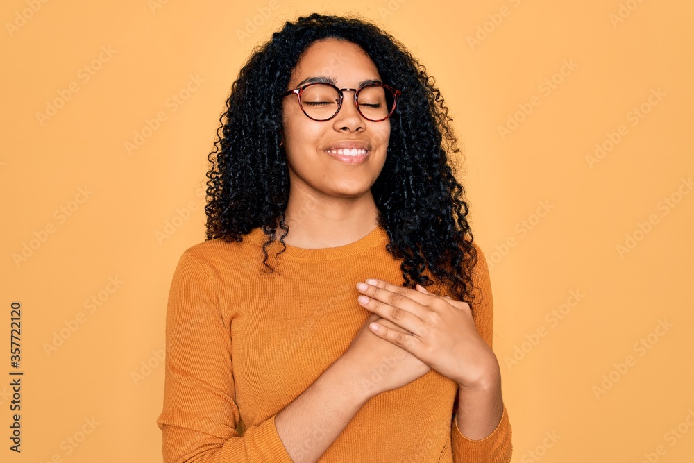 Young african american woman wearing casual sweater and glasses over yellow background smiling with hands on chest with closed eyes and grateful gesture on face. Health concept. Young african american woman wearing casual sweater and glasses over yellow background smiling with hands on chest with closed eyes and grateful gesture on face. Health concept.