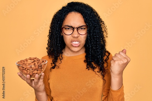 Young african american curly woman holding bowl with healthy almonds over yellow background annoyed and frustrated shouting with anger, crazy and yelling with raised hand, anger concept