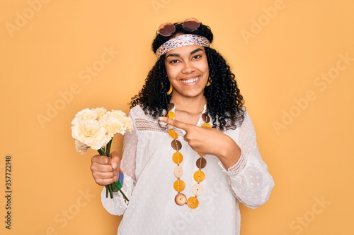 Young african american curly hippie woman wearing sunglasses holding bouquet of flowers very happy pointing with hand and finger
