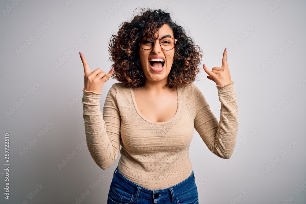 Young beautiful curly arab woman wearing casual t-shirt and glasses over white background shouting with crazy expression doing rock symbol with hands up. Music star. Heavy music concept. Young beautiful curly arab woman wearing casual t-shirt and glasses over white background shouting with crazy expression doing rock symbol with hands up. Music star. Heavy music concept.