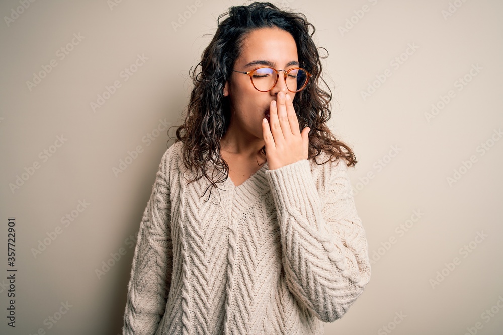 Beautiful woman with curly hair wearing casual sweater and glasses over white background bored yawning tired covering mouth with hand. Restless and sleepiness. Beautiful woman with curly hair wearing casual sweater and glasses over white background bored yawning tired covering mouth with hand. Restless and sleepiness.
