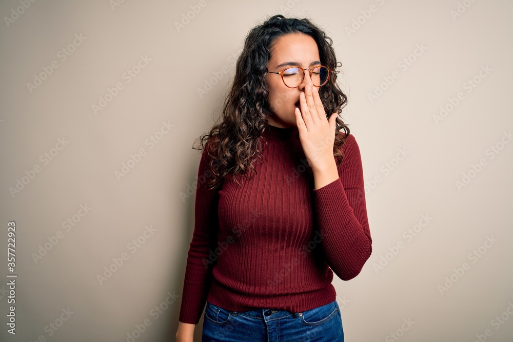 Beautiful woman with curly hair wearing casual sweater and glasses over white background bored yawning tired covering mouth with hand. Restless and sleepiness. Beautiful woman with curly hair wearing casual sweater and glasses over white background bored yawning tired covering mouth with hand. Restless and sleepiness.