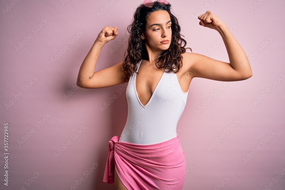 Beautiful woman with curly hair on vacation wearing white swimsuit over pink background showing arms muscles smiling proud. Fitness concept. Beautiful woman with curly hair on vacation wearing white swimsuit over pink background showing arms muscles smiling proud. Fitness concept.