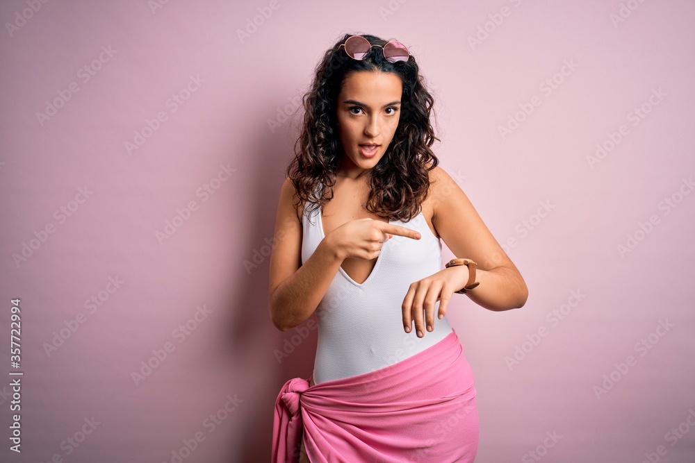 Beautiful woman with curly hair on vacation wearing white swimsuit over pink background In hurry pointing to watch time, impatience, upset and angry for deadline delay Beautiful woman with curly hair on vacation wearing white swimsuit over pink background In hurry pointing to watch time, impatience, upset and angry for deadline delay