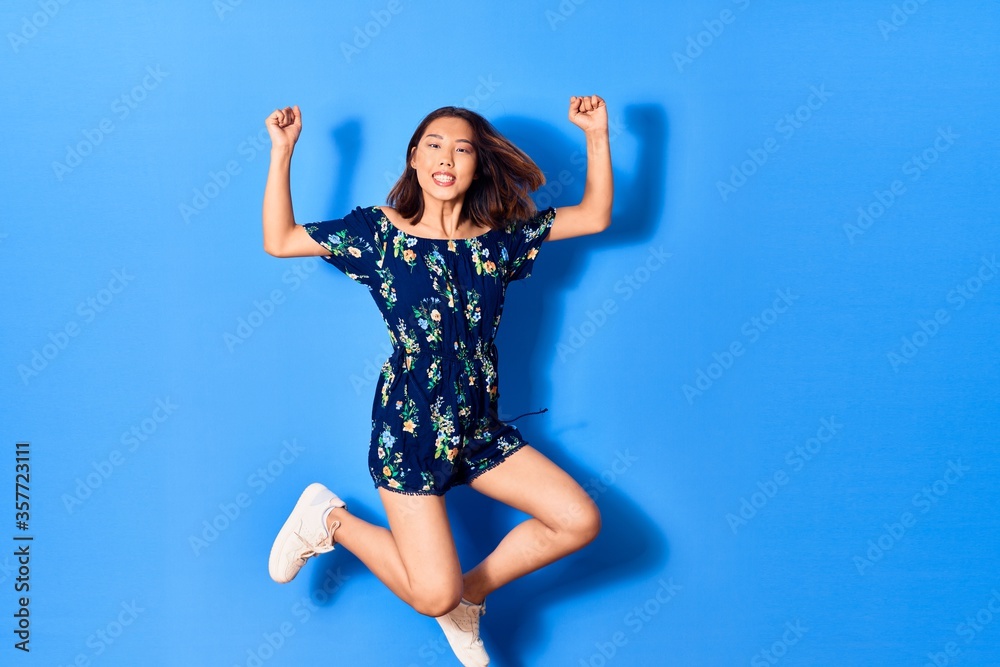 Naklejka premium Young beautiful chinese woman wearing casual clothes smiling happy. Jumping with smile on face doing strong geture over isolated blue background