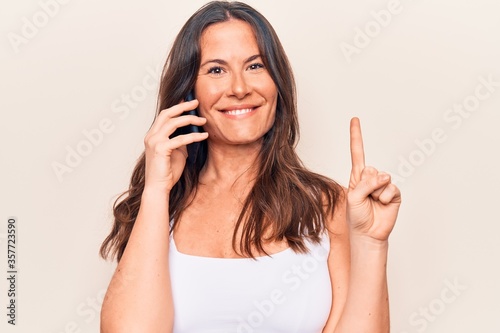 Beautiful brunette woman having conversation talking on the smartphone over white background smiling with an idea or question pointing finger with happy face, number one