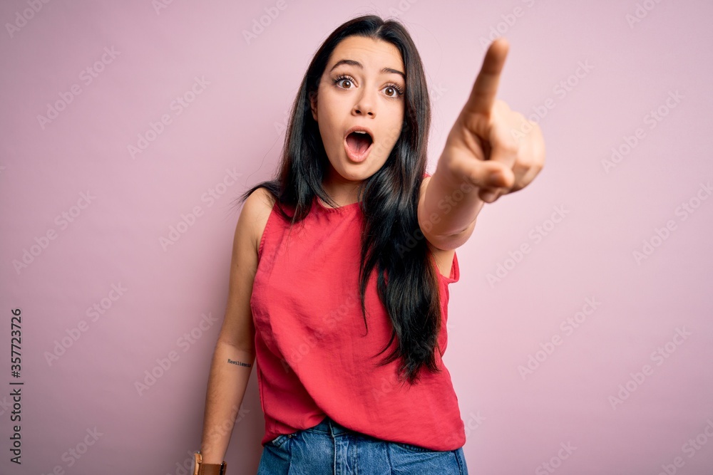Young brunette woman wearing casual summer shirt over pink isolated background Pointing with finger surprised ahead, open mouth amazed expression, something on the front