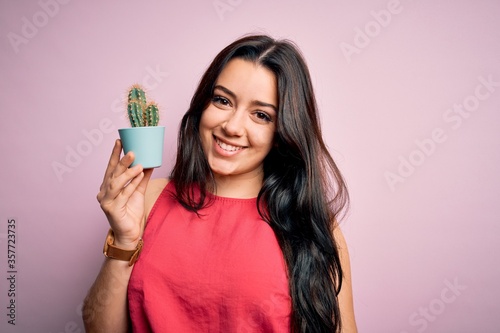 Young brunette woman holding succulent cactus plant over pink background with a happy face standing and smiling with a confident smile showing teeth